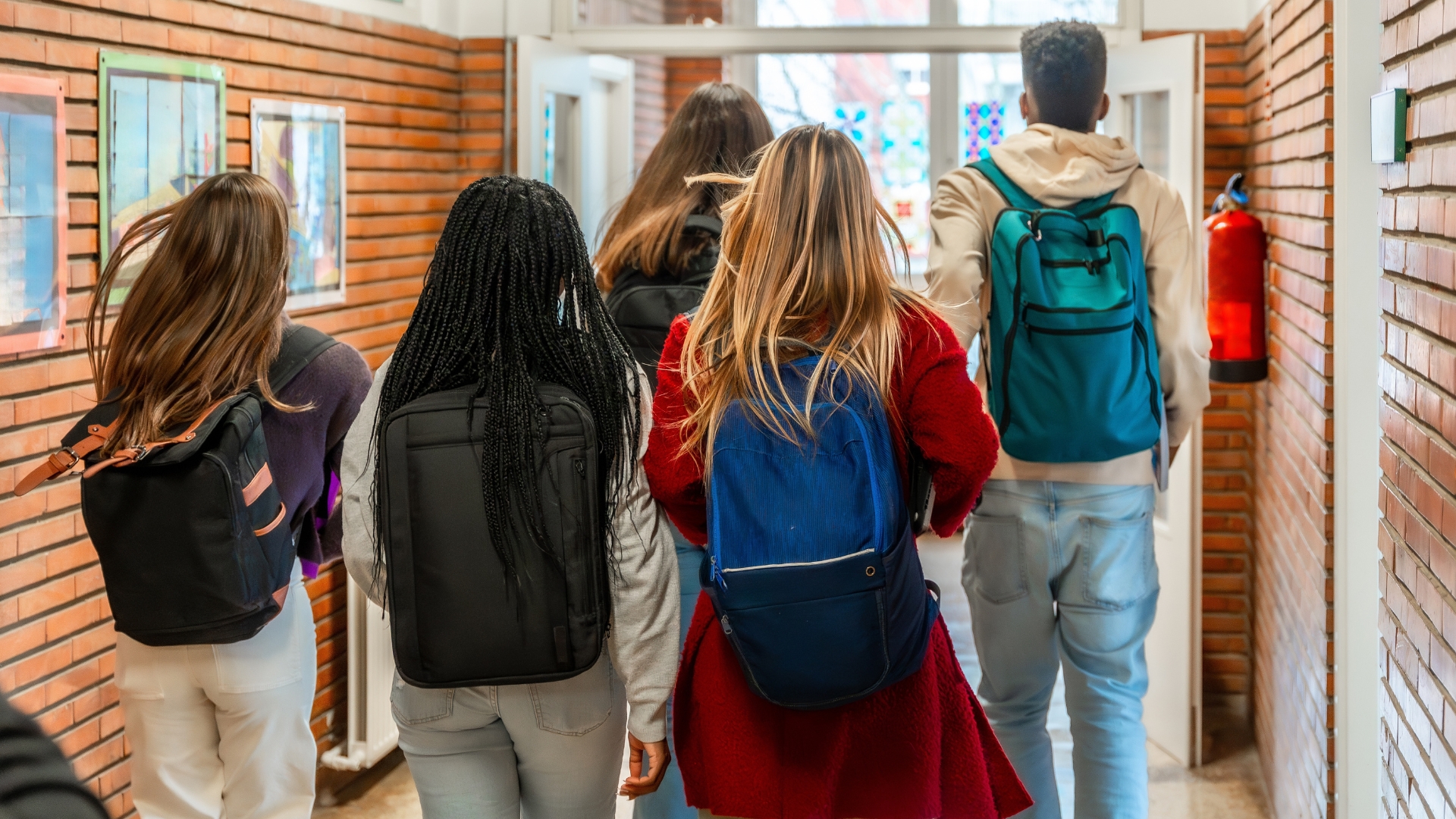 A rear view of five students carrying backpacks walking down a school hallway lined with brick walls toward an open door.